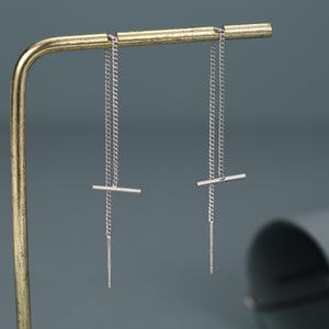 Könnte beinhalten: Ein Paar silberfarbene Durchziehohrringe mit einem zarten, feinen Kettendesign. Jeder Ohrring besteht aus einem dünnen Metallstab, der an einer Kette befestigt ist, die durch das Ohr gezogen wird und in einem geraden, schlanken Stift endet.