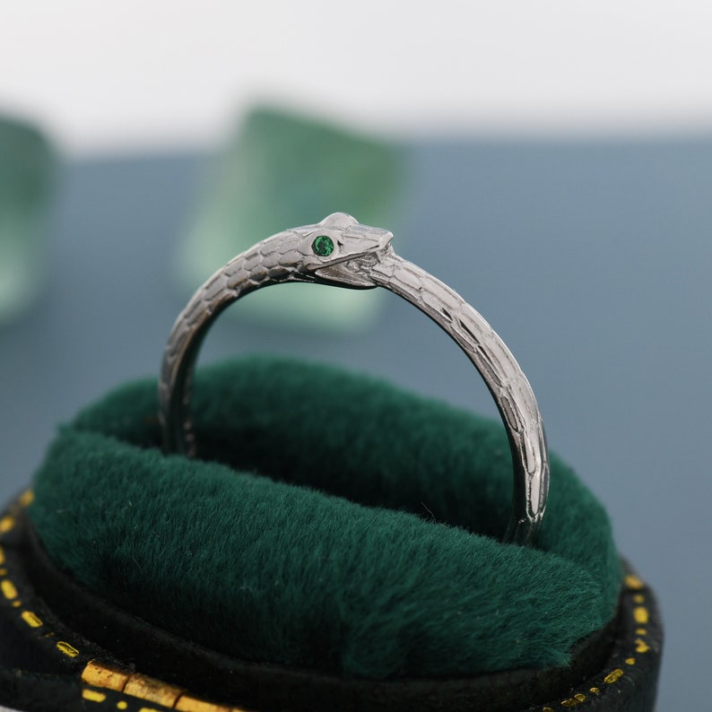 May include: A silver snake ring with a green gemstone eye. The ring is in a green velvet ring box.