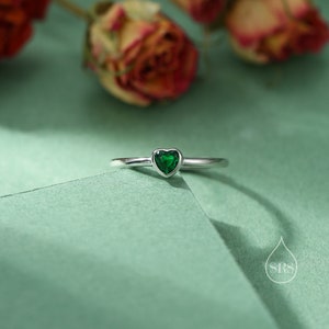 Può includere: Un anello d'argento con una pietra preziosa verde a forma di cuore. L'anello è su una superficie verde.