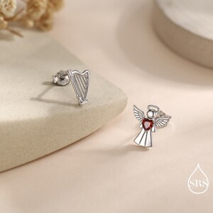 May include: Silver-toned stud earrings. One earring is a harp, the other is an angel with a red heart-shaped center. The earrings are on a neutral background.