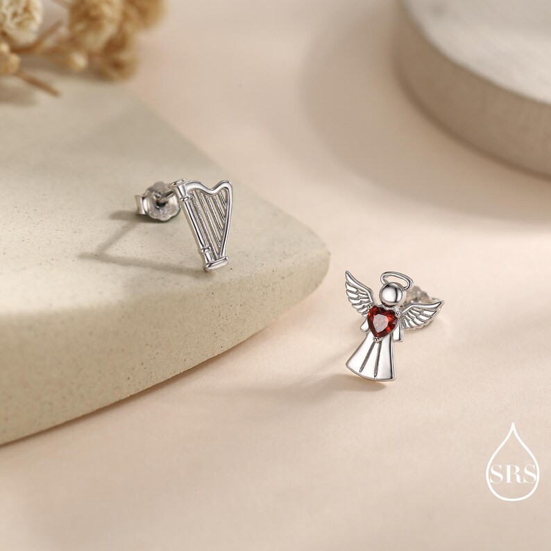 May include: Silver-toned stud earrings. One earring is a harp, the other is an angel with a red heart-shaped center. The earrings are on a neutral background.