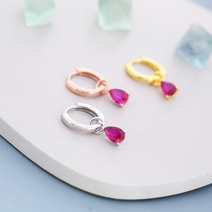 May include: Three pairs of hoop earrings with a dangling teardrop-shaped red gemstone. The hoops are available in gold, rose gold, and silver.