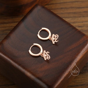 May include: A pair of rose gold hoop earrings with a dangling eye of Horus charm. The earrings are on a wooden surface.