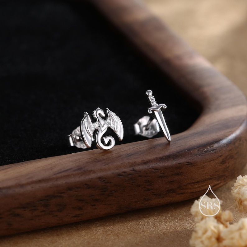 Peut inclure: Une paire de boucles d'oreilles clous argent&eacute;es. L'une repr&eacute;sente un dragon aux ailes d&eacute;ploy&eacute;es, l'autre une &eacute;p&eacute;e. Les boucles d'oreilles sont pr&eacute;sent&eacute;es sur une surface sombre dans un plateau en bois.