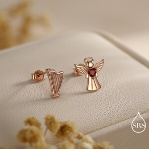 May include: Rose gold stud earrings. One earring is a harp, the other is an angel with a red heart-shaped gemstone. The earrings are displayed on a cream-colored surface with a blurred background.