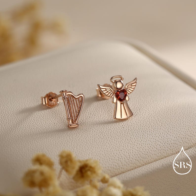 May include: Rose gold stud earrings. One earring is a harp, the other is an angel with a red heart-shaped gemstone. The earrings are displayed on a cream-colored surface with a blurred background.