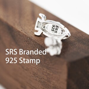 May include: Close-up of a silver earring with a rocket ship design. The earring has a 925 stamp and the letters "SRS" on the back.
