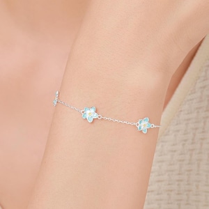 May include: A silver bracelet with three light blue and yellow flower charms. The bracelet is displayed on a wrist, highlighting the floral design and chain details. The flowers have a light blue enamel and yellow centre. The bracelet is a delicate piece of jewellery.