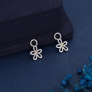 May include: Two silver flower charms with gold centres. Each charm has a small loop at the top. The charms are displayed on a dark blue surface, with a dark blue background.