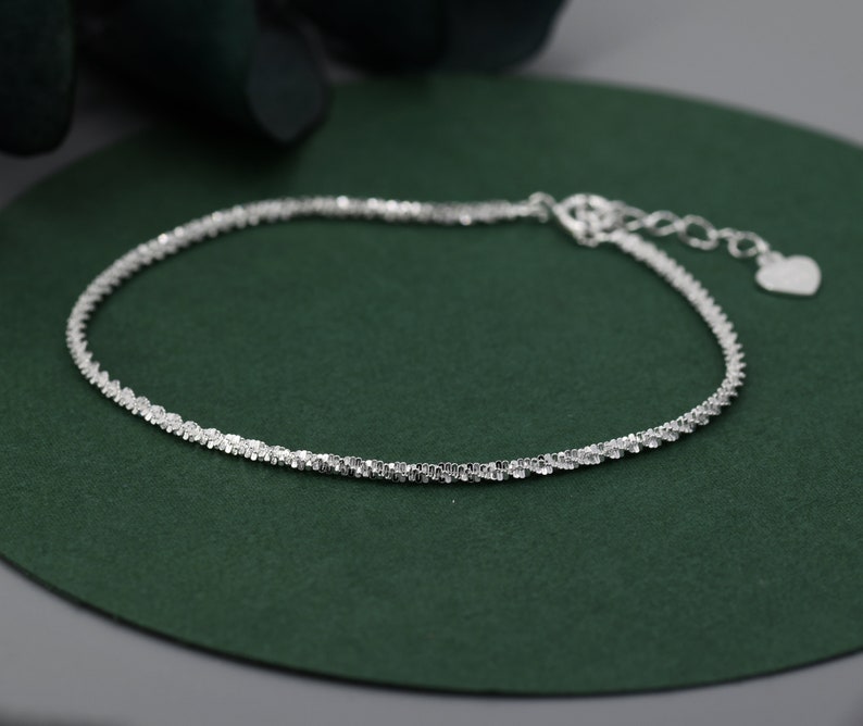 Puede incluir: Un brazalete de cadena de plata con un dije en forma de coraz&oacute;n. La cadena es de dise&ntilde;o delicado y trenzado.