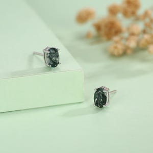 Puede incluir: Un par de pendientes de botón con piedras preciosas ovaladas de color verde oscuro engastadas en metal plateado. Los pendientes se muestran sobre una superficie verde claro, con un fondo suave y borroso.
