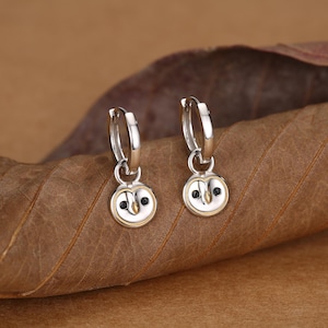 Può includere: Un paio di orecchini a cerchio in argento con ciondoli circolari a forma di viso di gufo. Ogni ciondolo presenta una finitura in argento lucido, un piccolo becco color oro e due minuscoli occhi neri circolari. Gli orecchini hanno una chiusura a scatto.