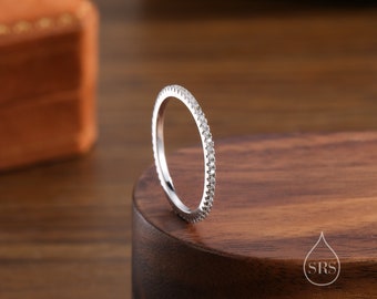 May include: A silver ring with a band of small, sparkling stones. The ring is set on a dark wooden surface, with a blurred orange box in the background. The ring is a simple, elegant design.