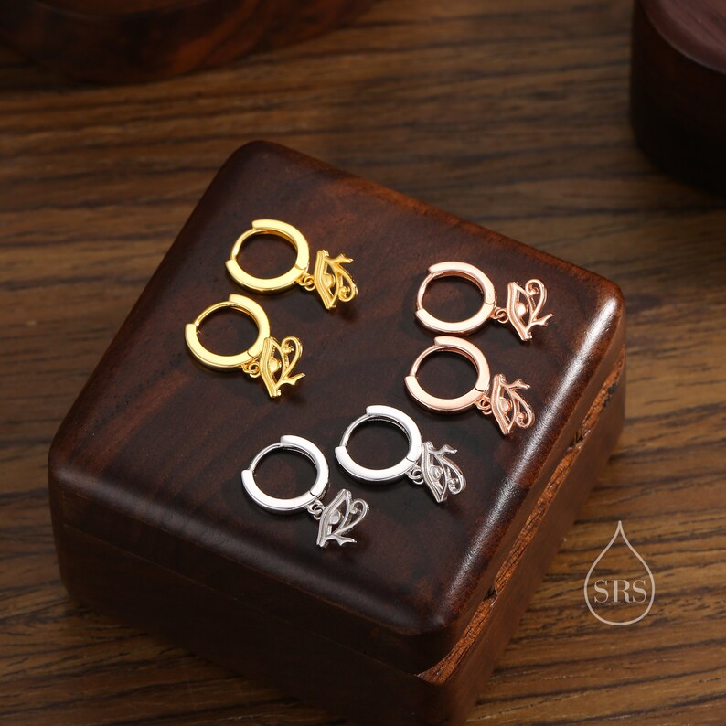 May include: Four pairs of hoop earrings with a dangling charm in the shape of an eye. The earrings are displayed in a wooden box. The earrings are available in gold, rose gold, and silver.