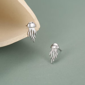 Può includere: Un paio di orecchini a lobo color argento a forma di medusa. Ogni orecchino presenta una parte superiore liscia e arrotondata con diversi tentacoli sottili e ondulati che scendono verso il basso, creando un design marino molto dettagliato.