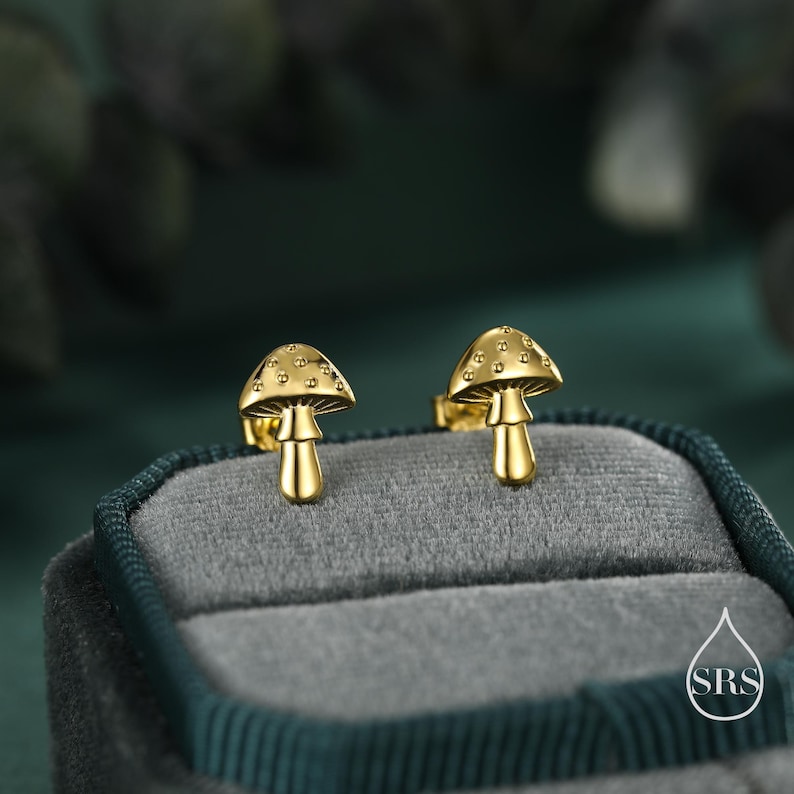 May include: A pair of gold-colored mushroom stud earrings. The earrings are displayed in a gray velvet jewelry box. The mushroom caps have a textured surface with small, round details. The stems are smooth and cylindrical.
