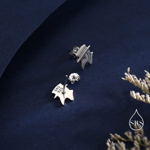 May include: A pair of silver-colored stud earrings. One earring is a stylized design of a plague doctor with a hat and beak. The other earring is a small, silver-colored leaf shape. The earrings are on a dark blue background.