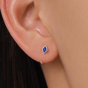 May include: A silver stud earring featuring a blue, diamond-shaped gemstone. The earring is set in a silver bezel and is worn on an ear. The skin tone is light, and the background is blurred.