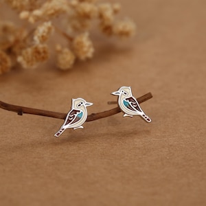Op de afbeelding: Een paar zilveren oorknopjes in de vorm van kookaburra-vogels. Elke oorbel is voorzien van emaille details in crème, bruin en een klein accent van helderblauw op de vleugel. De vogels zijn in profiel afgebeeld met zilveren omlijningen.