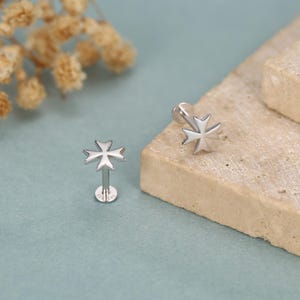May include: Two silver-colored stud earrings, each featuring a Maltese cross design. One earring is upright, showing the post and backing, while the other is angled. The earrings are set against a light blue and beige backdrop.