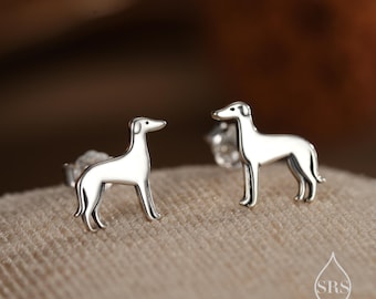 May include: A pair of silver-colored stud earrings featuring a Greyhound dog design. The earrings are detailed with the dog's silhouette, showcasing its slender body and long legs. The earrings are set on a beige surface.