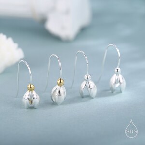 May include: Four silver-colored earrings with a flower bud design. Each earring has a curved hook and a small gold-colored bead. The earrings are displayed on a light blue background.