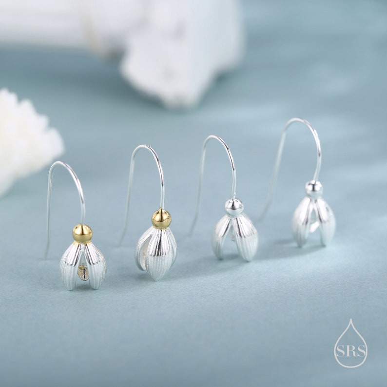 May include: Four silver-colored earrings with a flower bud design. Each earring has a curved hook and a small gold-colored bead. The earrings are displayed on a light blue background.