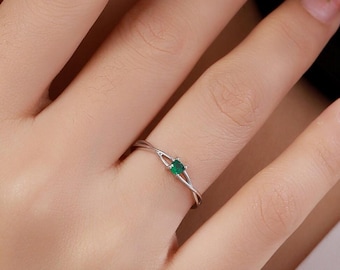 Puede incluir: Un delicado anillo de plata con una pequeña esmeralda de corte cuadrado. La banda tiene un diseño retorcido, añadiendo un toque de elegancia. El anillo se muestra en un dedo, resaltando sus finos detalles y su estilo minimalista.