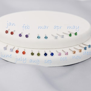 Puede incluir: Doce pares de peque&ntilde;os pendientes de bot&oacute;n con piedras preciosas de diferentes colores, dispuestos en c&iacute;rculo sobre un expositor blanco. Los meses del a&ntilde;o est&aacute;n escritos en azul debajo de cada par de pendientes.