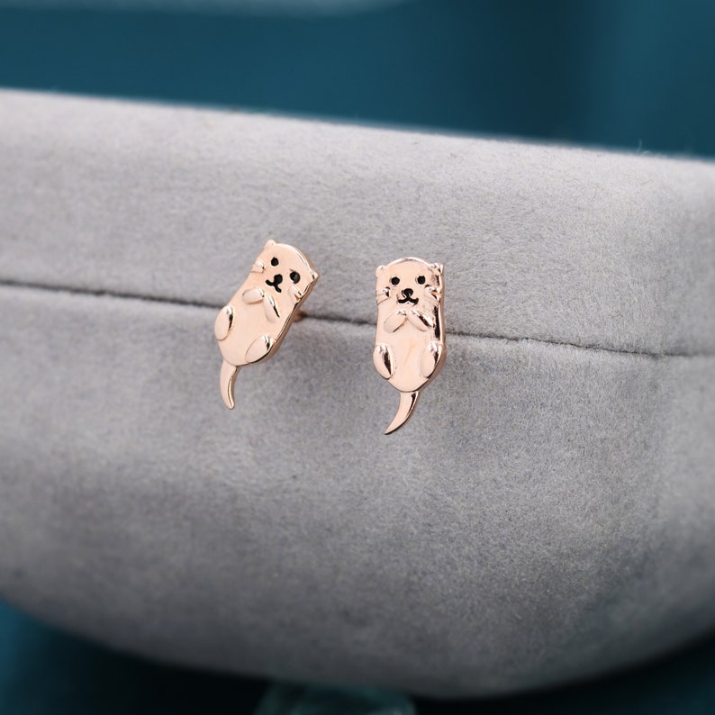 May include: A pair of gold-toned earrings featuring a cute otter design. The otters are facing forward with their paws raised, as if they are waving.