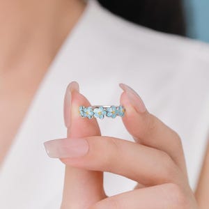 Peut inclure: Une bague argentée ornée d'une bande continue de petites fleurs en émail bleu clair. Chaque fleur possède cinq pétales et un minuscule centre jaune, formant un motif floral délicat qui fait tout le tour de l'anneau en métal.