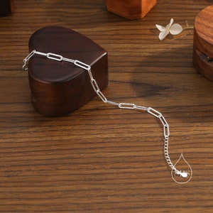 May include: A silver chain bracelet with a delicate, textured, rectangular link design. The bracelet is displayed on a wooden surface with a heart-shaped box.
