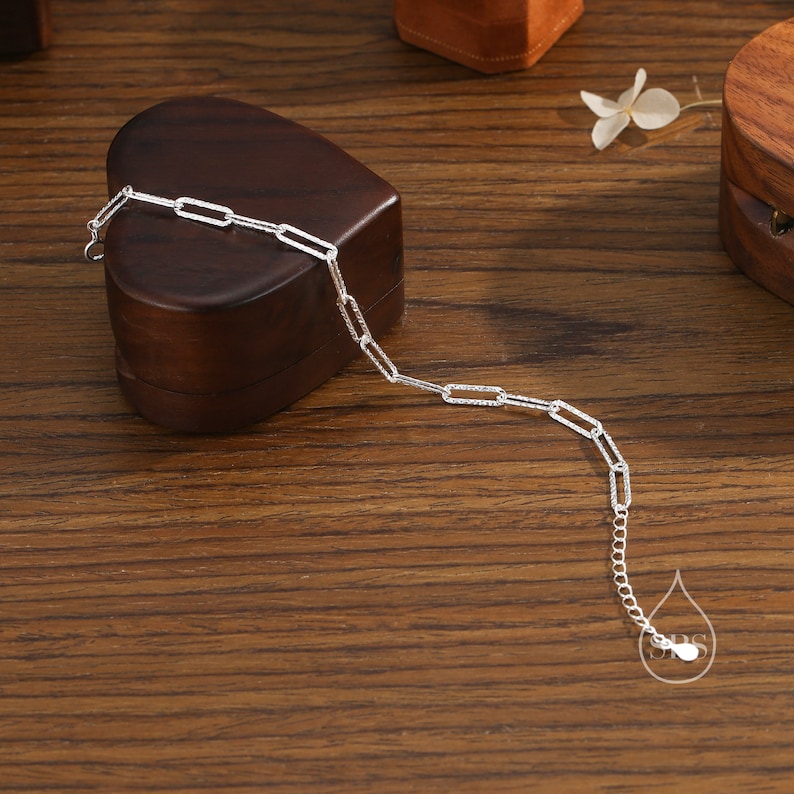May include: A silver chain bracelet with a delicate, textured, rectangular link design. The bracelet is displayed on a wooden surface with a heart-shaped box.