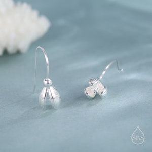 May include: Silver-toned floral drop earrings. Each earring features a delicate flower design with detailed petals and a curved hook for pierced ears. The earrings are displayed against a soft blue background.
