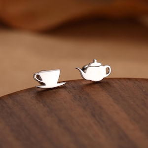 Könnte beinhalten: Silberne Ohrstecker mit einem Teetassen- und Teekannen-Design. Die Teetasse hat einen Henkel und steht auf einer kleinen Untertasse. Die Teekanne hat einen gebogenen Ausguss und einen kleinen Deckel. Die Ohrringe liegen auf einer Holzoberfläche.