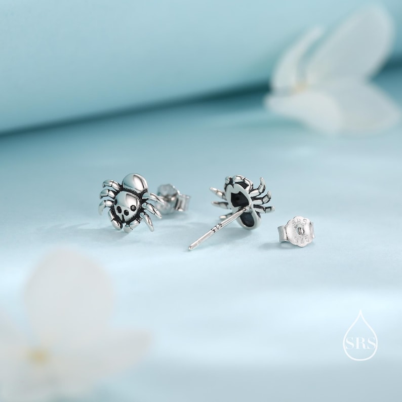 May include: A pair of silver spider stud earrings. The spiders have black eyes and are detailed with intricate carvings. The earrings are on a white background.