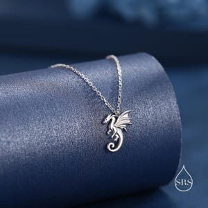May include: A silver-toned necklace featuring a small dragon pendant with outstretched wings and a curled tail. The pendant is suspended from a delicate, thin silver-toned chain. A small logo with the letters SRS inside a water drop shape is visible.