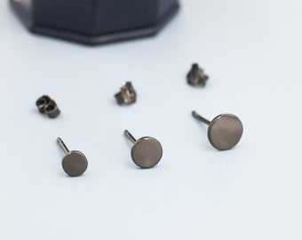 May include: Three pairs of black stud earrings with round metal tops. The earrings are arranged on a light blue surface.