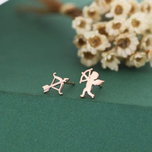 May include: Rose gold stud earrings featuring an arrow and bow design, and a cupid figure. The earrings are displayed on a green surface with white flowers in the background. The earrings are small and delicate.