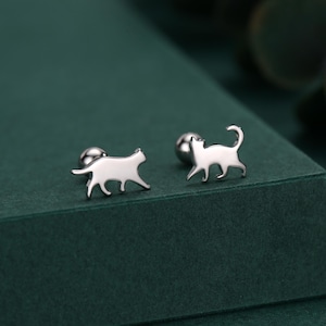 Könnte beinhalten: Ein Paar silberfarbene Ohrstecker in Katzenform. Jeder Ohrring hat eine kleine, stilisierte Katzensilhouette mit gebogenem Schwanz und einem Stift mit rundem Rücken. Die Ohrringe werden auf einer dunkelgrünen Oberfläche präsentiert.