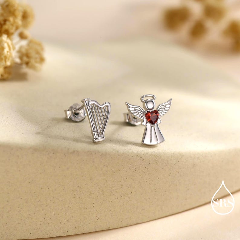 May include: A pair of silver stud earrings. One earring is a harp, the other is an angel with a red heart-shaped gemstone. The angel has wings and a halo. The earrings are on a neutral-toned surface.