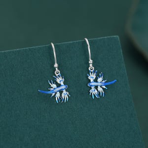 Può includere: Un paio di orecchini pendenti in argento con un design a lumaca di mare blu. Gli orecchini hanno un aspetto dettagliato e stilizzato con accenti di smalto blu e bianco. Gli orecchini sono sospesi a un semplice gancio d'argento.