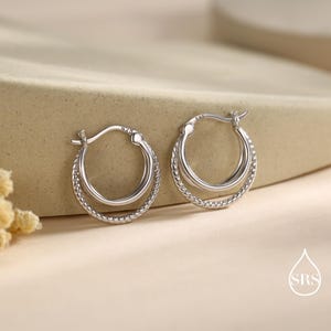 Peut inclure: Une paire de boucles d'oreilles créoles argentées. Chaque boucle d'oreille présente un design à double couche, avec un cercle intérieur lisse et un cercle extérieur texturé et torsadé. Les boucles d'oreilles sont présentées sur une surface neutre.