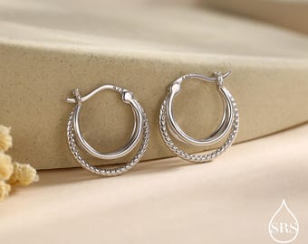 May include: A pair of silver hoop earrings. Each earring features a double-layered design, with a smooth inner hoop and a textured, twisted outer hoop. The earrings are displayed on a neutral-toned surface, showcasing their reflective qualities.