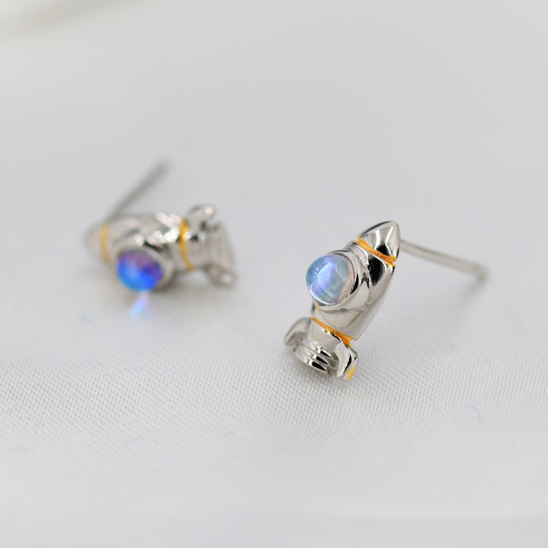 May include: A pair of silver rocket ship earrings with iridescent blue gemstones. The rocket ships have gold accents.