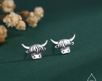May include: A pair of silver earrings featuring a Highland cow design. The earrings are small and delicate, with a detailed depiction of the cow's head and horns.