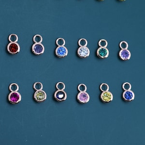 Peut inclure: Une collection de pendentifs dorés, chacun orné d'une petite pierre précieuse colorée. Les pierres précieuses sont de différentes couleurs, dont le rouge, le bleu, le vert et le violet. Les pendentifs sont disposés sur un fond bleu canard foncé.