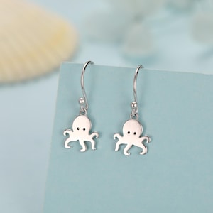 May include: A pair of silver octopus earrings. Each earring features a small, stylized octopus charm with a simple design. The earrings have a curved hook for pierced ears and a small ball detail.