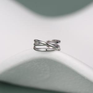 May include: A polished silver-toned ring featuring a multi-band design with three distinct strands that cross over each other at the centre, creating an open, layered appearance with a smooth, reflective metallic finish throughout the entire piece.