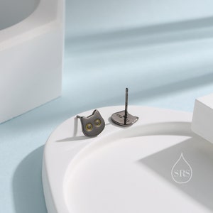 Peut inclure: Une paire de boucles d'oreilles clous en forme de chat noir avec des accents dor&eacute;s. Les boucles d'oreilles sont sur une surface blanche.